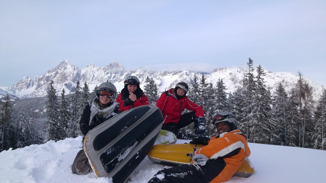 Eine Gruppe von vier Personen sitzt im Schnee und hält Schlitten. Im Hintergrund sind schneebedeckte Berge und Tannen zu sehen. | © OCT