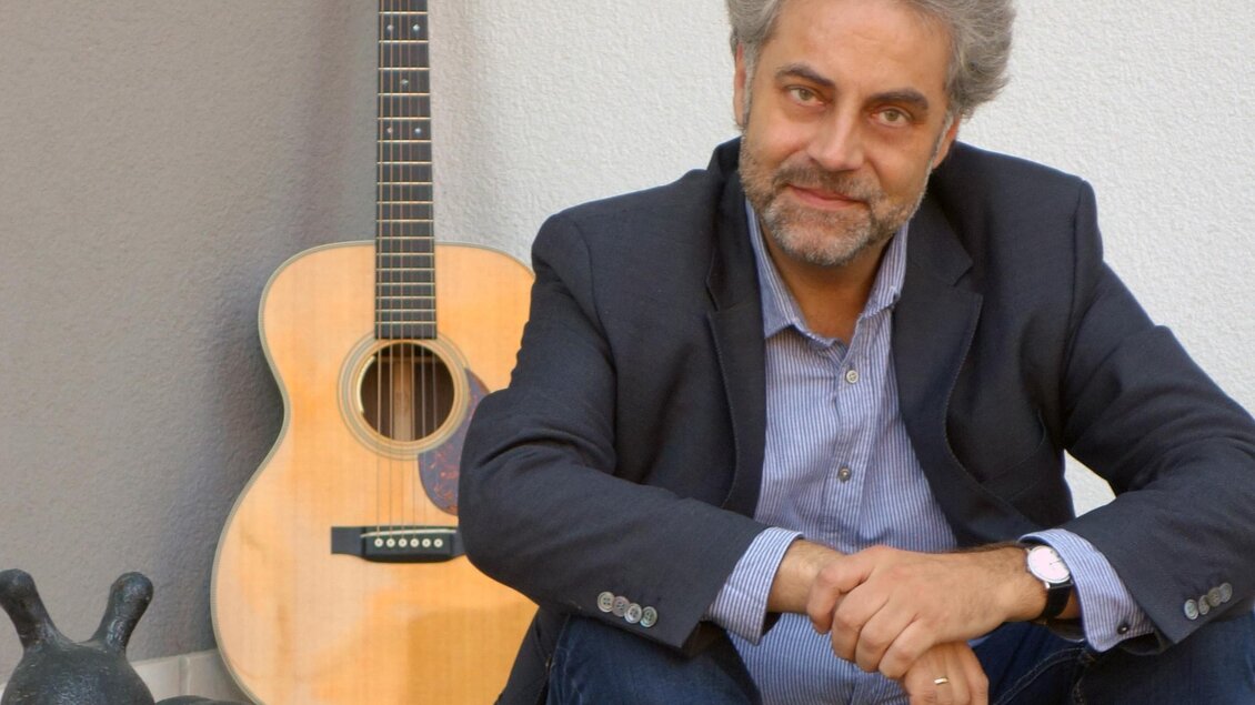 Ein Mann sitzt lächelnd auf dem Boden neben einer Gitarre. Im Hintergrund ist eine Skulptur einer Schnecke sichtbar.