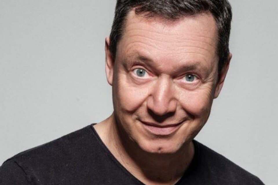 Ein lächelnder Mann mit kurzem, dunklem Haar trägt ein schwarzes T-Shirt. Der Hintergrund ist neutral und grau. | © congress Schladming