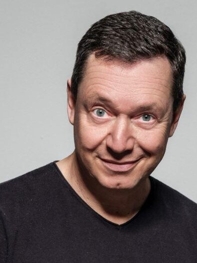 Ein lächelnder Mann mit kurzem, dunklem Haar trägt ein schwarzes T-Shirt. Der Hintergrund ist neutral und grau. | © congress Schladming