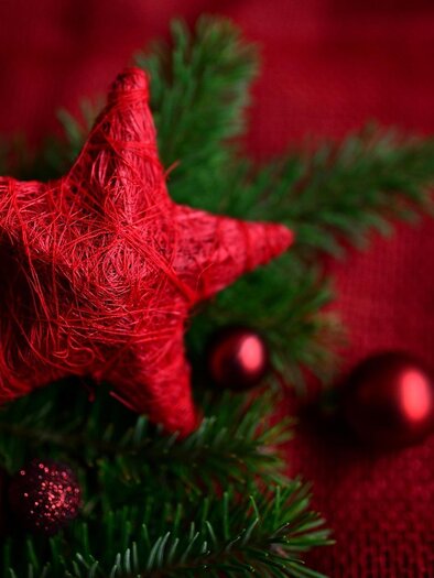 A red star made of yarn lies on green branches. In the background, red Christmas ornaments can be seen. | © pixabay.com
