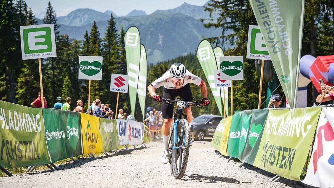 Mountainbiker sprintet ins Ziel der Alpentour in Schladming-Dachstein, Zuschauer jubeln entlang der Strecke mit grünen und gelben Bannern. | © Sportograf