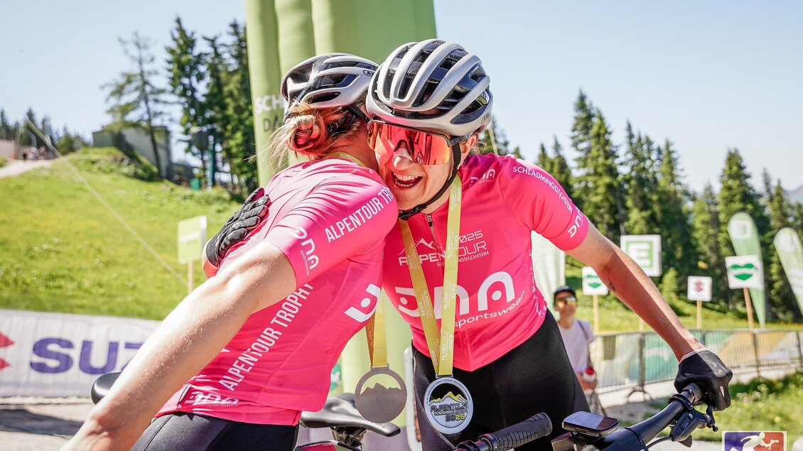 Zwei Radfahrerinnen mit pinken Trikots umarmen sich jubelnd im Zielbereich, Medaillen um den Hals.
 | © Sportograf