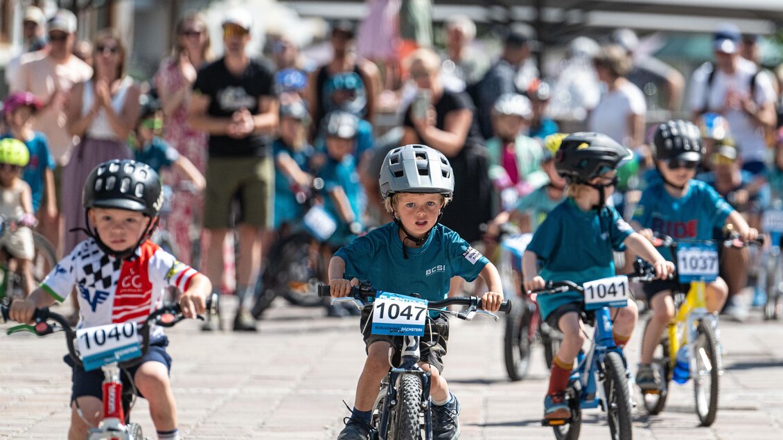 Kleine Kinder mit Helmen fahren bei einem Radrennen auf Laufrädern, Zuschauer klatschen im Hintergrund. | © Alpentour Trophy