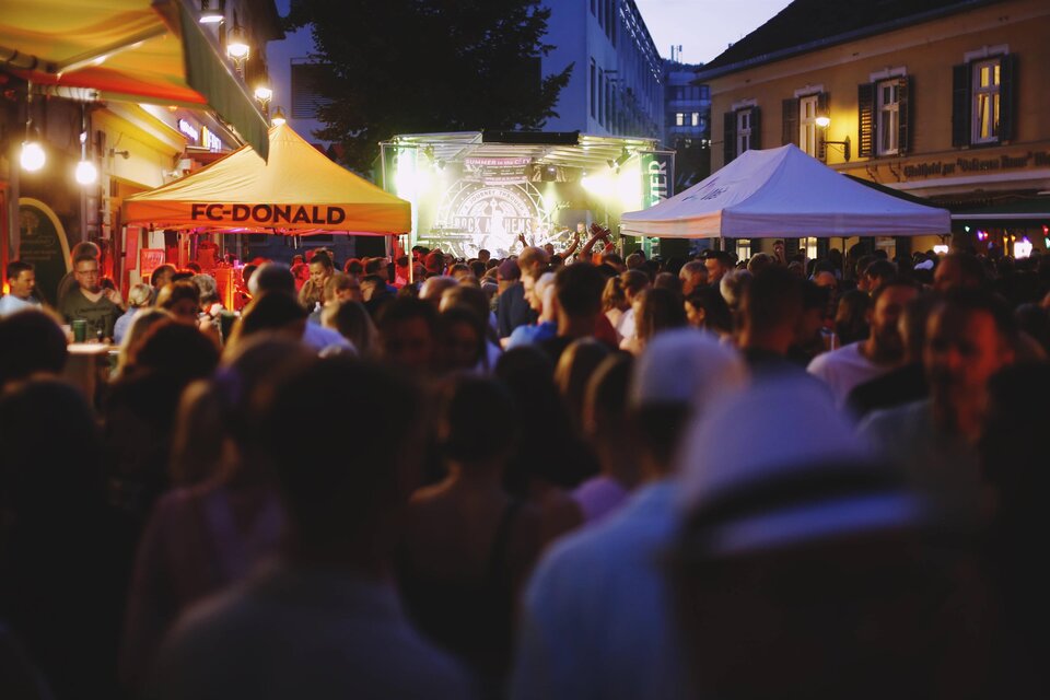 Eine belebte Veranstaltung bei Einbruch der Dunkelheit mit vielen Menschen. Es gibt mehrere Stände, darunter einen mit der Aufschrift "FC-DONALD". | © Tauser