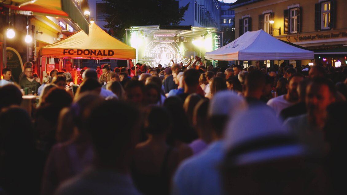 Eine belebte Veranstaltung bei Einbruch der Dunkelheit mit vielen Menschen. Es gibt mehrere Stände, darunter einen mit der Aufschrift "FC-DONALD". | © Tauser