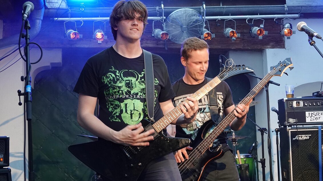 Zwei Musiker spielen live auf der Bühne. Der Gitarrist trägt ein schwarzes T-Shirt und spielt ein elektrisches Gitarre, während der Bassist im Hintergrund steht. | © Heran