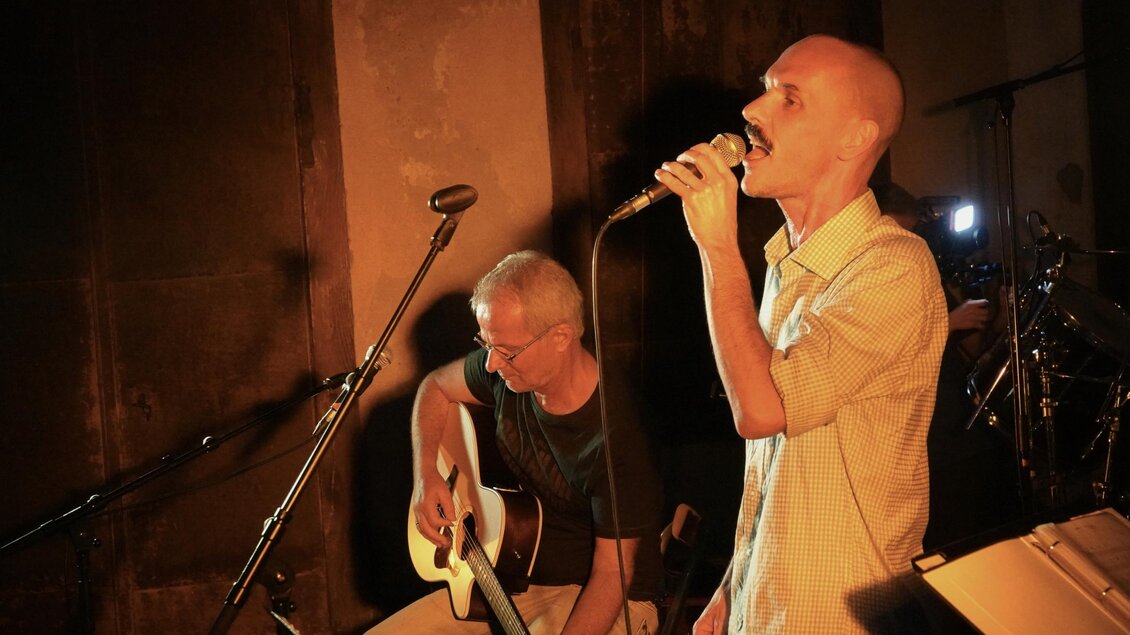 Zwei Musiker treten auf einer kleinen Bühne auf. Einer singt ins Mikrofon, während der andere Gitarre spielt. | © Heran