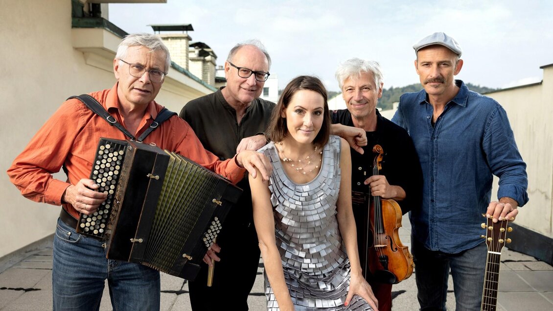 Eine Gruppe von fünf Musikern steht draußen und posiert für das Foto. Sie tragen verschiedene Instrumente und lächeln freundlich in die Kamera. | © ANIADA A NOAR