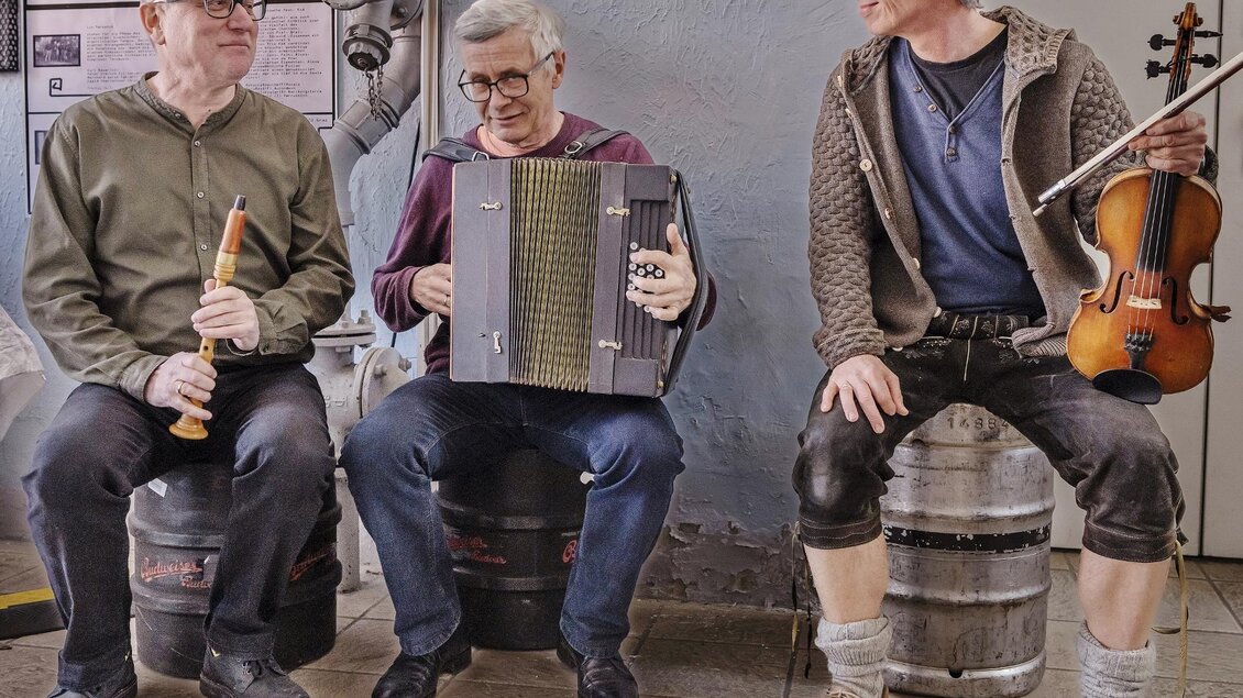 Drei Männer sitzen auf Fässern und spielen gemeinsam Musik. Einer spielt Flöte, einer ein Akkordeon und der dritte eine Geige. | © Kranzlbinder
