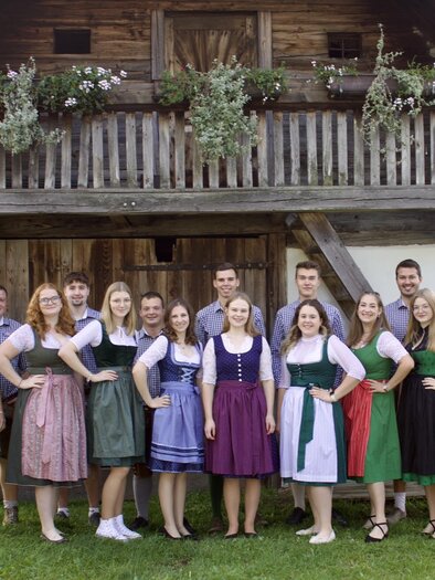 A group of people in traditional costume stands in front of a rustic half-timbered house. The men and women are smiling and posing together in a beautiful rural setting. | © Landjugend Stubenberg