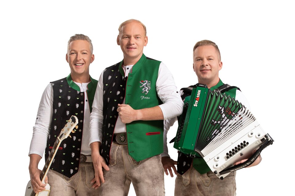 Drei Musiker stehen in Trachtenkleidung und lächeln in die Kamera. Einer spielt ein Akkordeon, während die anderen Gitarre und Gesang bereit halten. | © DIE EDLSEER/Fotograf Karl Schrotter