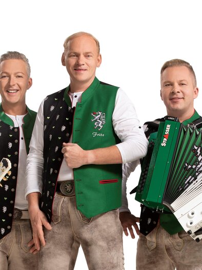 Three musicians are wearing traditional costumes and smiling at the camera. One is playing an accordion while the others are ready with a guitar and vocals. | © DIE EDLSEER/Fotograf Karl Schrotter