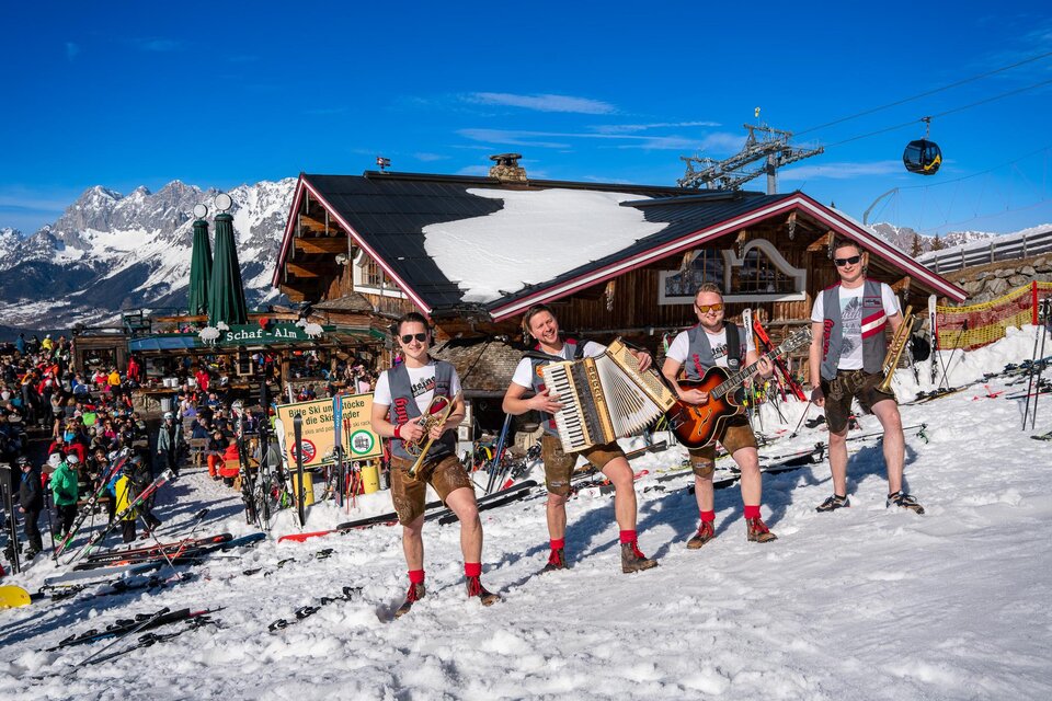 Eine fröhliche Gruppe von Musikern in traditioneller Kleidung spielt in einer schneebedeckten Berglandschaft. Im Hintergrund sind ein Skigebiet und viele Menschen zu sehen. | © Mario Egger