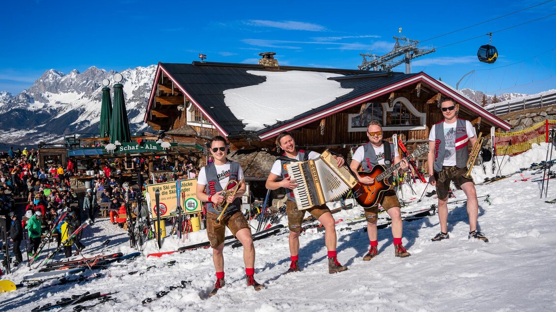 Eine fröhliche Gruppe von Musikern in traditioneller Kleidung spielt in einer schneebedeckten Berglandschaft. Im Hintergrund sind ein Skigebiet und viele Menschen zu sehen. | © Mario Egger