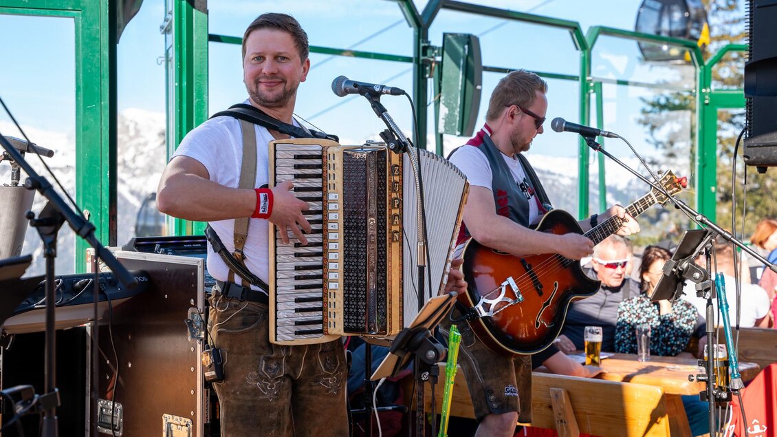Zwei Musiker treten in einer Berghütte auf. Einer spielt das Akkordeon, der andere die Gitarre, während die Gäste in der Sonne genießen. | © Mario Egger