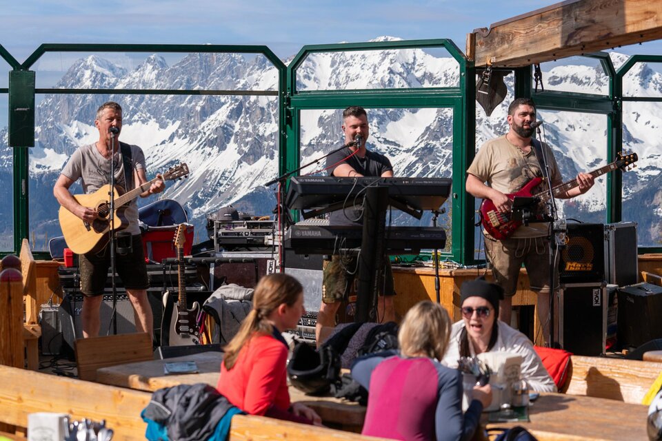 Eine Live-Band spielt in einer alpinen Umgebung mit schneebedeckten Bergen im Hintergrund. Menschen sitzen gemütlich und genießen die Musik in einem Außenbereich. | © Mario Egger