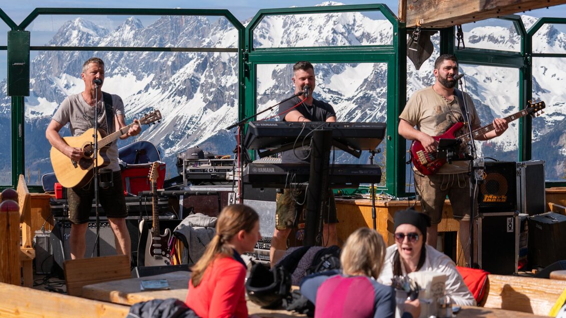 Eine Live-Band spielt in einer alpinen Umgebung mit schneebedeckten Bergen im Hintergrund. Menschen sitzen gemütlich und genießen die Musik in einem Außenbereich. | © Mario Egger