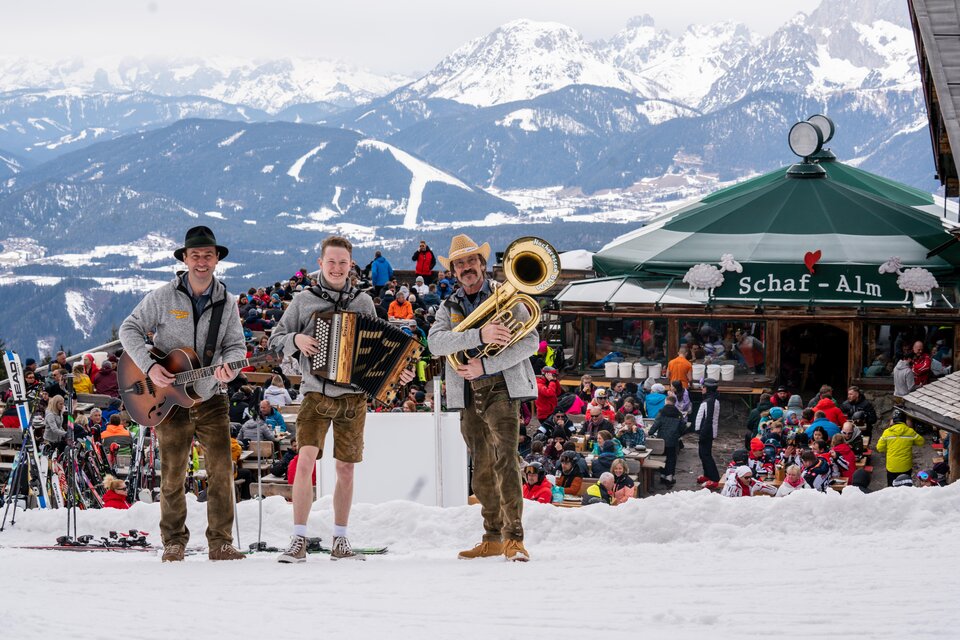 Drei Musiker stehen im Schnee vor einer Berghütte und spielen traditionelle Musik. Im Hintergrund sind viele Menschen und beeindruckende Berge zu sehen. | © „Hochgrössen Buam"