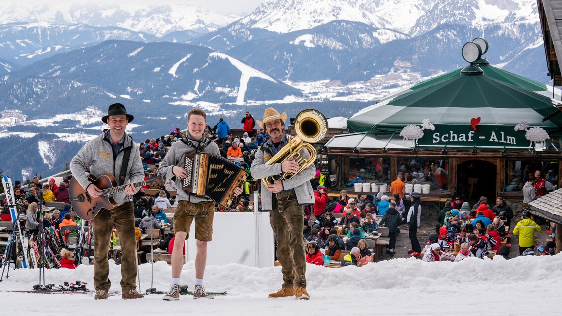 Drei Musiker stehen im Schnee vor einer Berghütte und spielen traditionelle Musik. Im Hintergrund sind viele Menschen und beeindruckende Berge zu sehen. | © „Hochgrössen Buam"