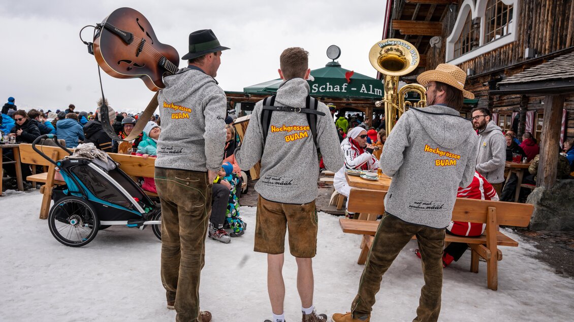 Drei Musiker stehen mit Instrumenten in einer winterlichen Almhütte. Im Hintergrund sieht man viele Gäste, die sich amüsieren und die prominente Umgebung genießen. | © Mario Egger