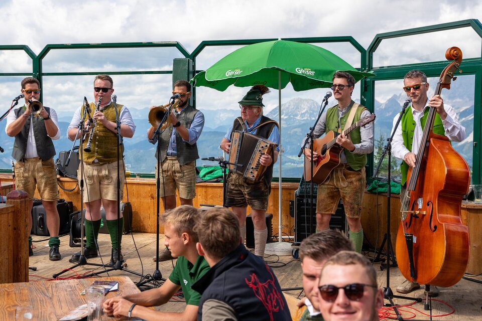 Eine Volksmusikkapelle spielt auf einer Bergterrasse. Im Hintergrund sind die Alpen und ein grünes Sonnenschirm zu sehen. | © Mario Egger