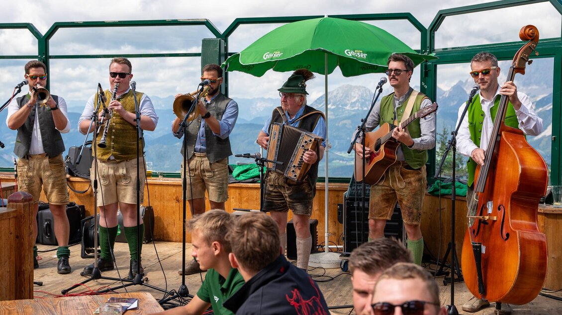 Eine Volksmusikkapelle spielt auf einer Bergterrasse. Im Hintergrund sind die Alpen und ein grünes Sonnenschirm zu sehen. | © Mario Egger