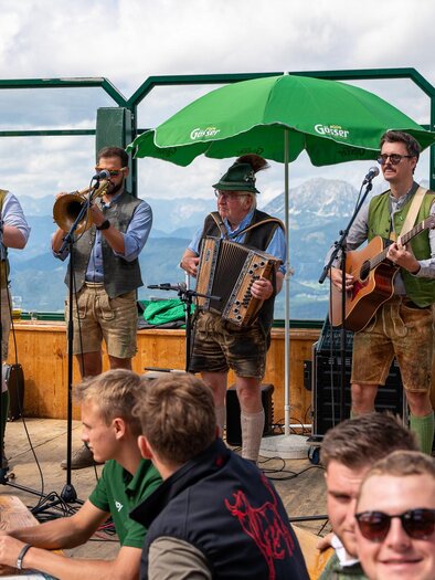 Eine Volksmusikkapelle spielt auf einer Bergterrasse. Im Hintergrund sind die Alpen und ein grünes Sonnenschirm zu sehen. | © Mario Egger