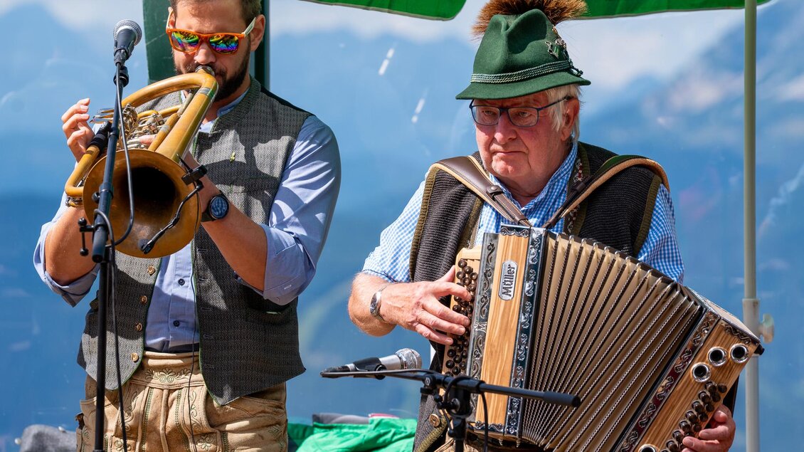Zwei Musiker in traditioneller Tracht spielen Trompete und Akkordeon. Im Hintergrund sieht man eine Berglandschaft und ein Sonnenschirm. | © Mario Egger