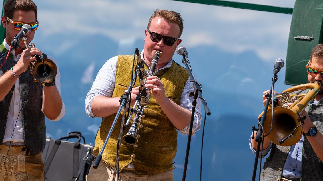Eine Gruppe von Musikern spielt auf einer Bühne in den Bergen. Sie tragen traditionelle Kleidung und spielen verschiedene Instrumente. | © Mario Egger
