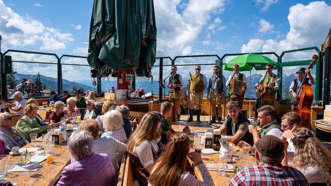 Eine lebendige Atmosphäre auf einer Bergterrasse mit einer Band, die traditionelle Musik spielt. Die Gäste genießen Essen und Getränke bei schönem Wetter und Blick auf die Berge. | © Mario Egger