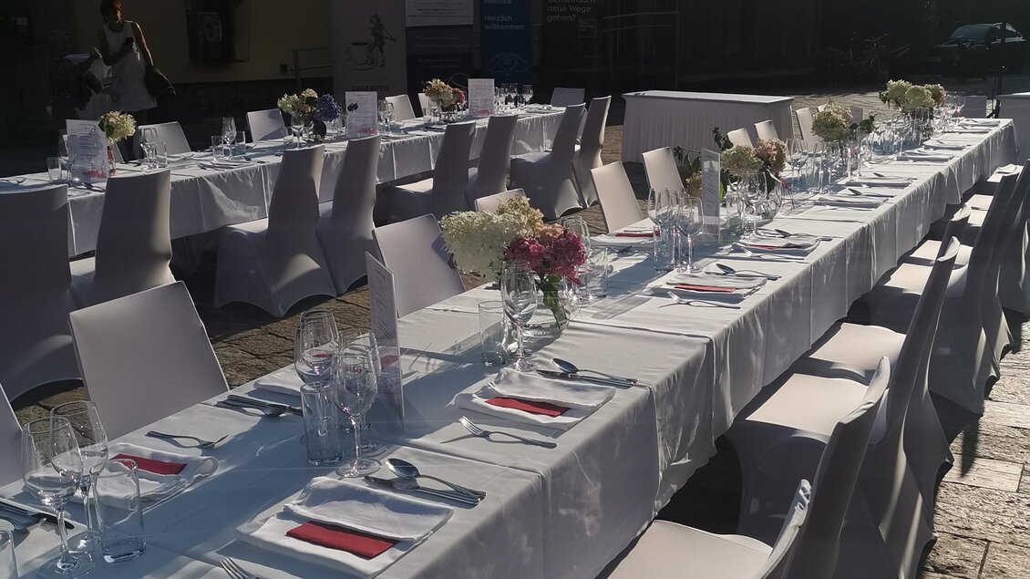 Ein elegantes gedecktes Tischarrangement im Freien, umgeben von Stühlen mit weißen Hussen. Der Tisch ist mit Blumen, Gläsern und Besteck dekoriert und bietet eine einladende Atmosphäre. | © CDM incoming - Fotostudio Augenblick