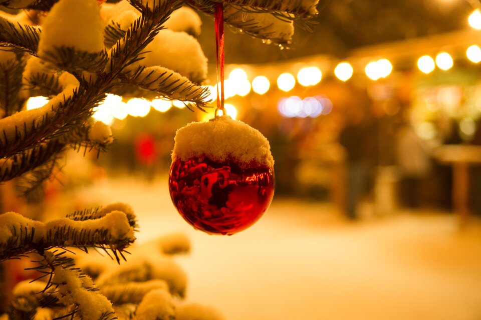 Eine rote Weihnachtskugel hängt an einem verschneiten Baum. Im Hintergrund sind die Lichter eines Weihnachtsmarktes zu sehen. | © Silvia Rastl