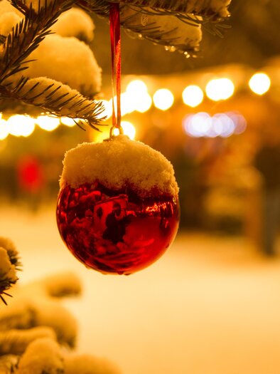 A red Christmas ornament hangs on a snow-covered tree. In the background, the lights of a Christmas market can be seen. | © Silvia Rastl