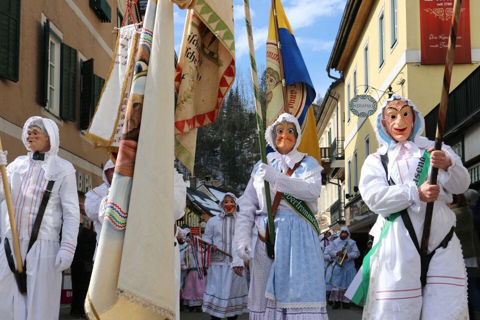Eine traditionelle Parade mit Menschen in weißen Kostümen und großen Masken. Die Teilnehmer halten Fahnen und marschieren durch eine malerische Straße. | © TVB Ausseerland - Salzkammergut_Zink