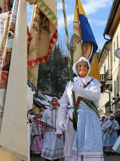 Eine traditionelle Parade mit Menschen in weißen Kostümen und großen Masken. Die Teilnehmer halten Fahnen und marschieren durch eine malerische Straße. | © TVB Ausseerland - Salzkammergut_Zink