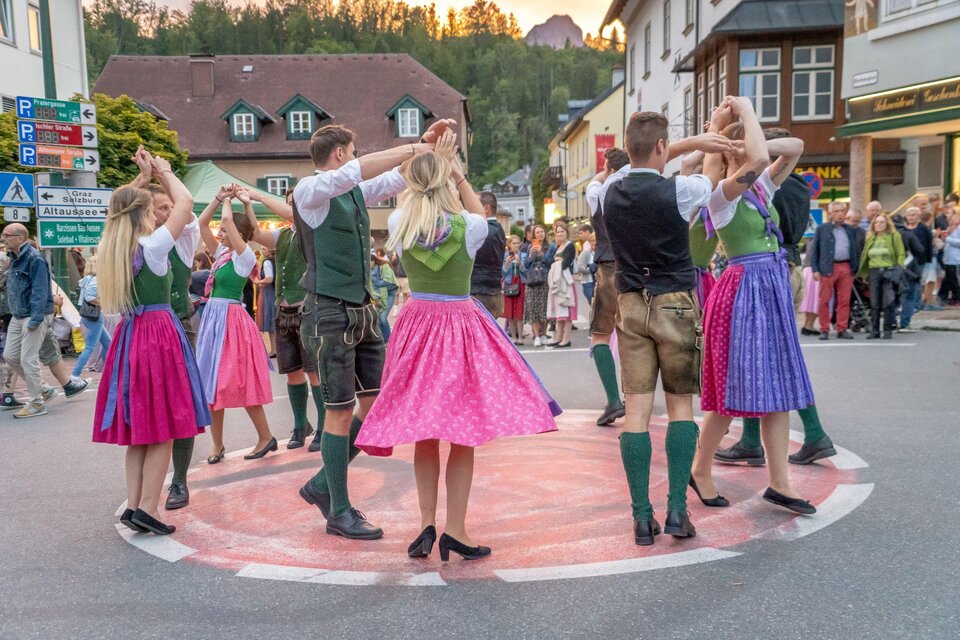 Eine Gruppe von Tänzern in traditionellen Trachten führt einen Volkstanz auf einer Straße vor einem Publikum auf. Die bunten Kleider und die festliche Stimmung schaffen eine fröhliche Atmosphäre. | © Christine Struz