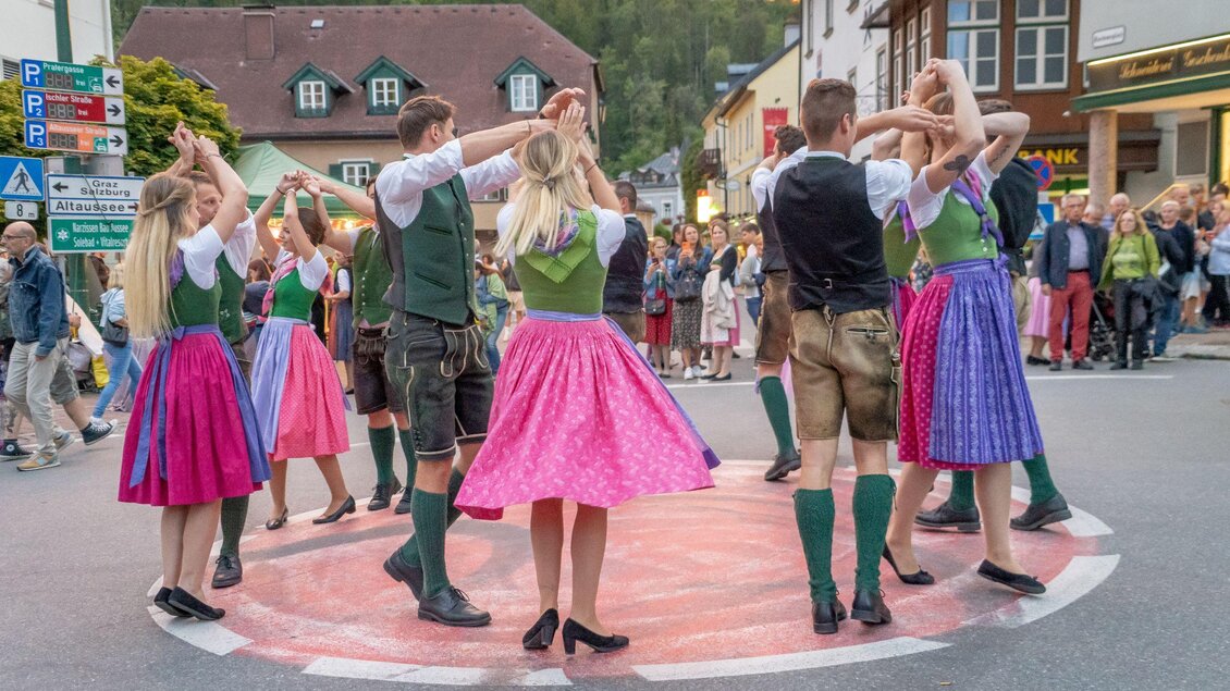 Eine Gruppe von Tänzern in traditionellen Trachten führt einen Volkstanz auf einer Straße vor einem Publikum auf. Die bunten Kleider und die festliche Stimmung schaffen eine fröhliche Atmosphäre. | © Christine Struz