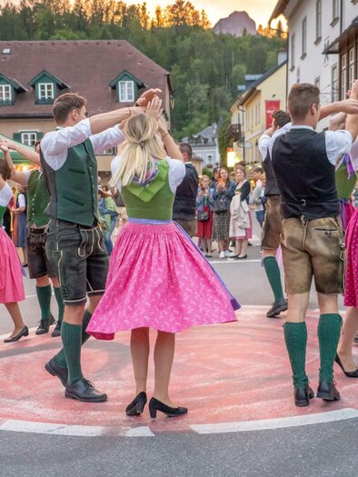 A group of dancers in traditional costumes performs a folk dance on a street in front of an audience. The colorful dresses and the festive mood create a cheerful atmosphere. | © Christine Struz