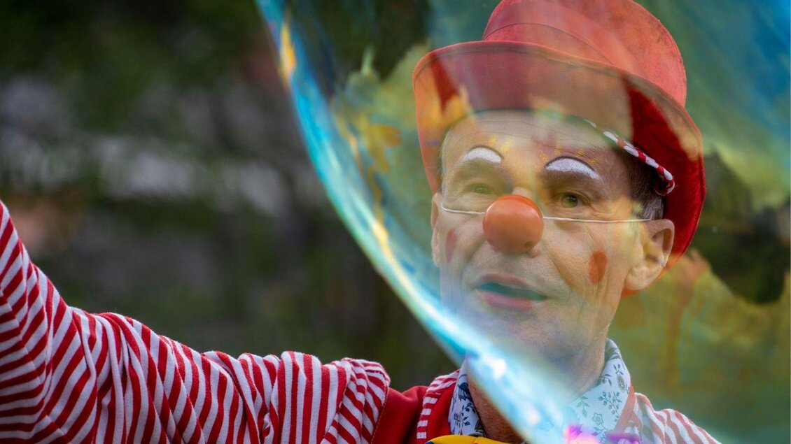 Ein Clown mit roten Streifen auf seinem Hemd und einem roten Hut bläst Seifenblasen. Sein Gesicht ist fröhlich bemalt und er lächelt. | © Christine Struz