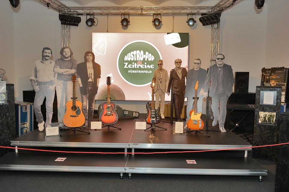 Eine Ausstellung mit mehreren Gitarren und lebensgroßen Pappaufstellern von Musikern. Im Hintergrund ist ein großes Banner mit dem Schriftzug "Austro-Pop Zeitreise". | © Austro-Pop Zeitreise