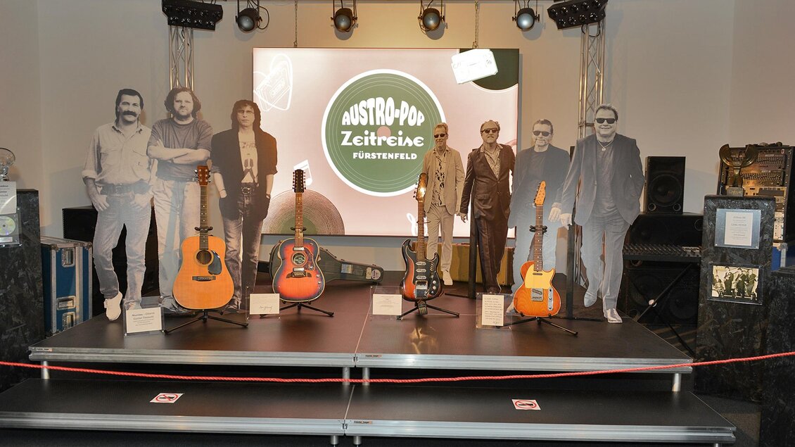 Eine Ausstellung mit mehreren Gitarren und lebensgroßen Pappaufstellern von Musikern. Im Hintergrund ist ein großes Banner mit dem Schriftzug "Austro-Pop Zeitreise". | © Austro-Pop Zeitreise