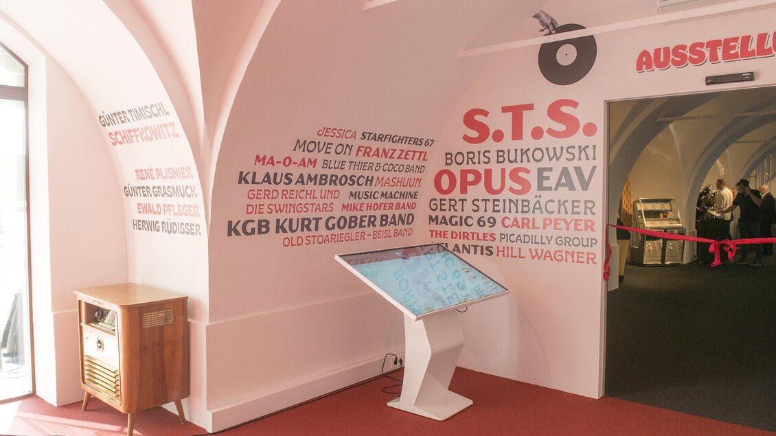 Ein Raum mit einer Ausstellung, in dem eine Wand mit verschiedenen Künstlernamen dekoriert ist. Im Vordergrund steht ein interaktiver Bildschirm auf einem weißen Tisch. | © Austro-Pop Zeitreise