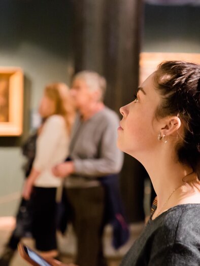 A young woman attentively examines a painting in a museum. In the background, other visitors can be seen exploring the exhibition. | © TV Südsteiermark - Adobe Stock