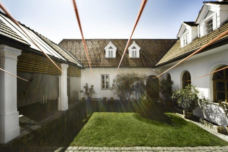 Ein ruhiger Innenhof mit grünem Rasen und einfachen Gebäuden. Die Sonne strahlt auf die Szene und erzeugt eine helle, einladende Atmosphäre. | © Galerie Gölles