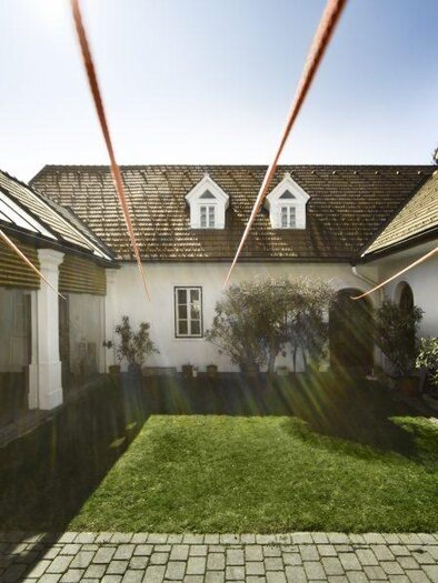 A quiet courtyard with green grass and simple buildings. The sun radiates on the scene, creating a bright, inviting atmosphere. | © Galerie Gölles