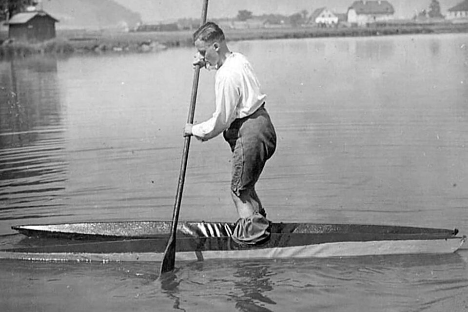 Ein Junge paddelt auf einem schmalen Boot auf ruhigem Wasser. Im Hintergrund sind Häuser und eine Landschaft zu sehen.