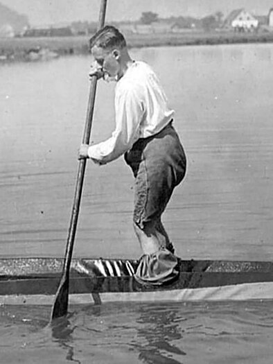 A boy paddles on a narrow boat on calm water. In the background, houses and a landscape can be seen.