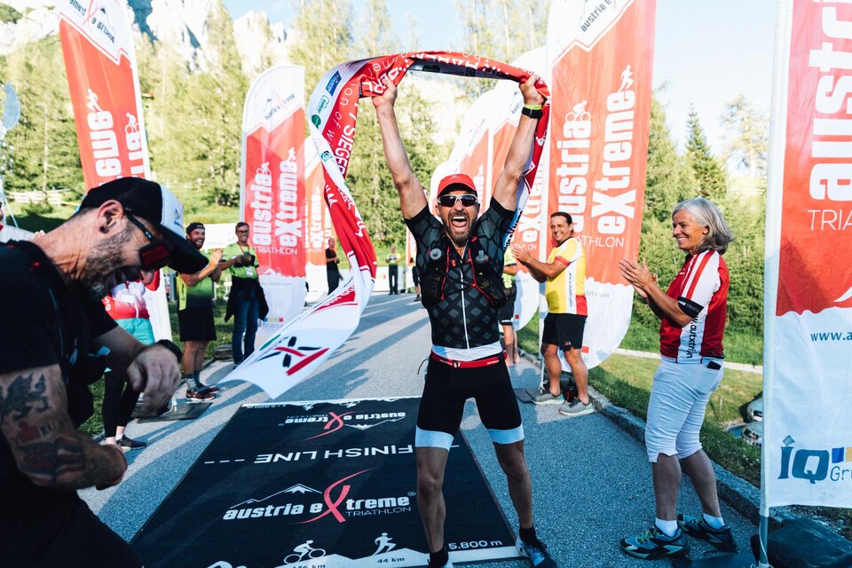 Ein begeisterter Sportler feiert den Zieleinlauf mit einer Flagge in der Hand. Im Hintergrund jubeln Zuschauer und weitere Teilnehmer. | © Austria eXtreme