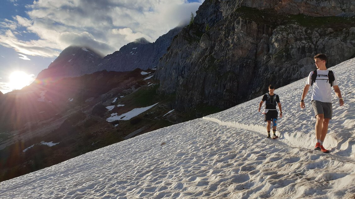 Zwei Wanderer gehen über einen schneebedeckten Bergpass. Die Sonne geht hinter den Bergen unter und beleuchtet die Szenerie. | © Austria eXtreme
